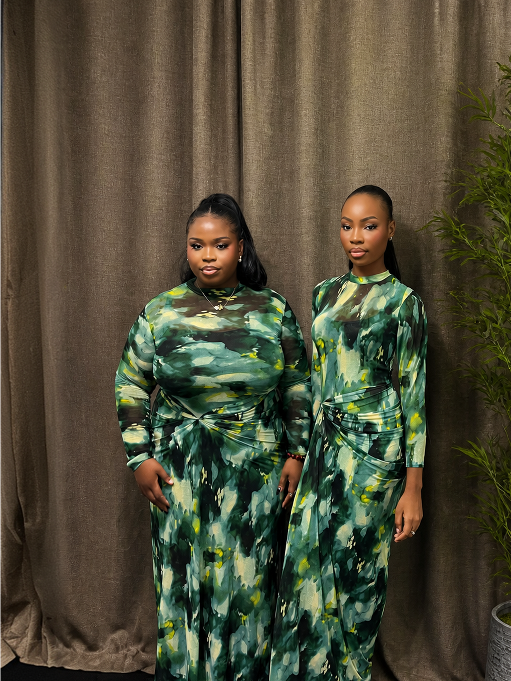 Two women wearing green tie-dye dresses standing against a brown curtain.
